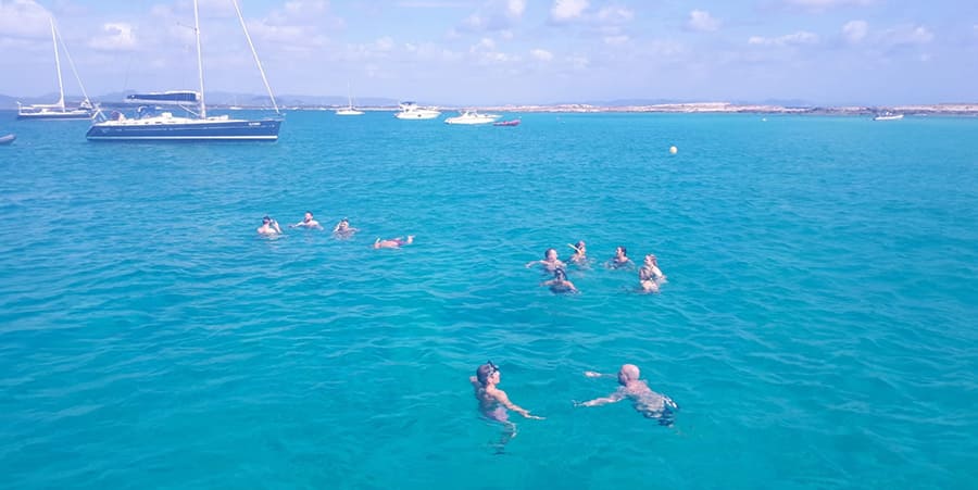 A group of people enjoy a swim in the ocean. There are boats in the distance. 