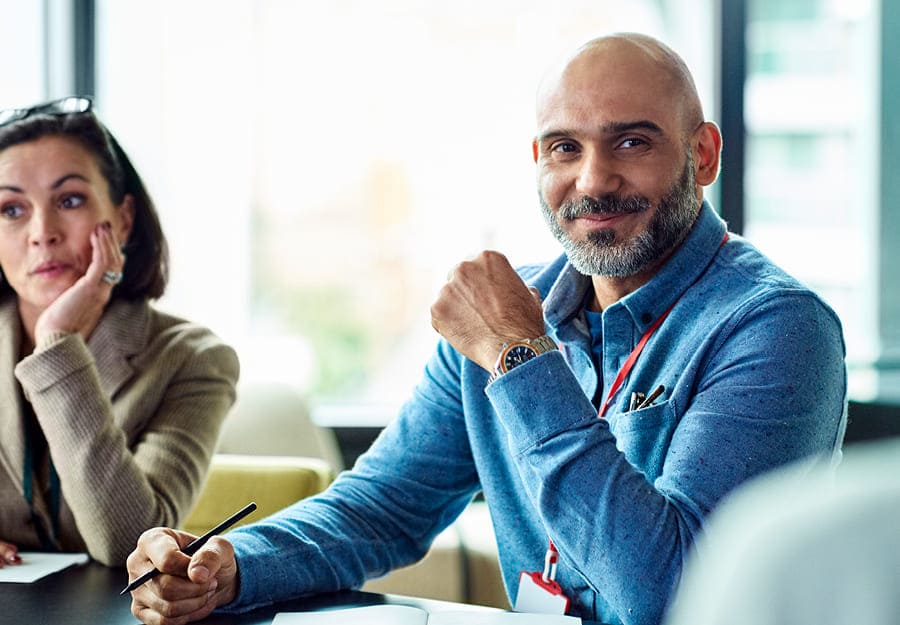 A professional setting with a man and woman sitting at a table. The man in focus wears a blue shirt. There's a notepad in front of him and he holds a pen in one hand. He's smiling.